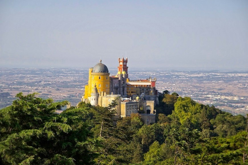 Colorful castle on a hilltop surrounded by trees with city view in the background.